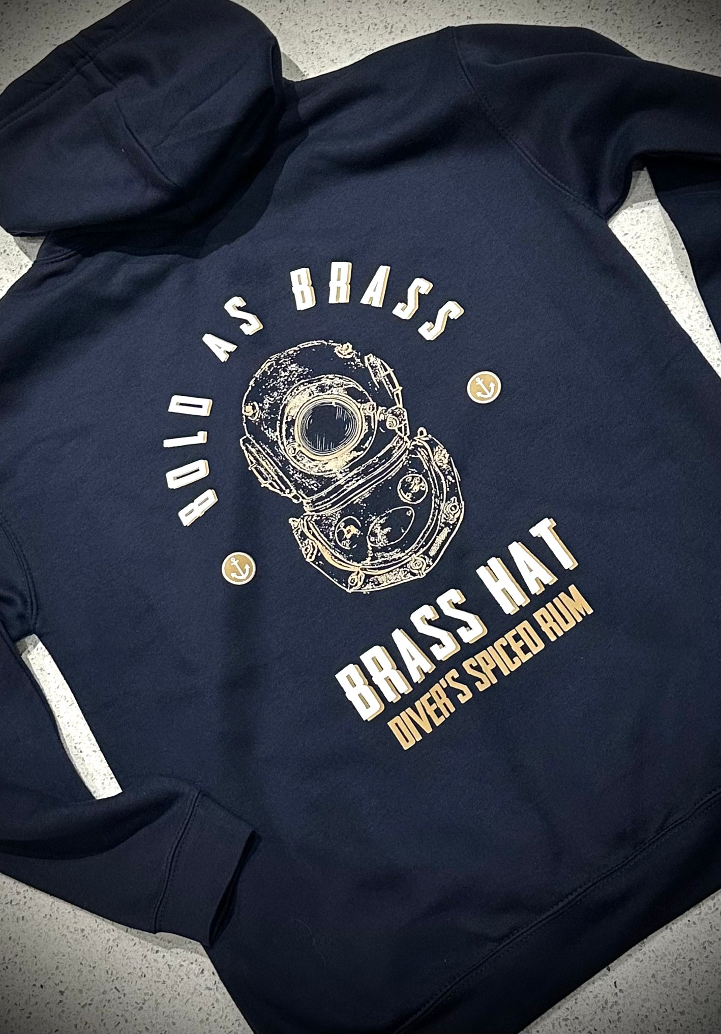 Back view of a navy blue hoodie in with bold as brass written on it,  a diving helmet logo, anchor motifs and Brass Hat Diver's Spiced Rum brand name