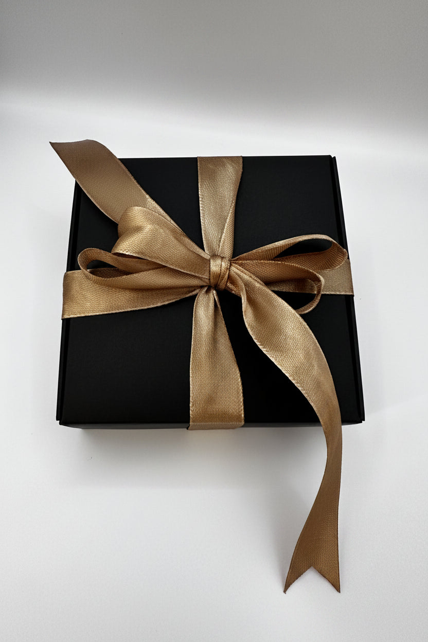 A square black gift box tied with a wide metallic gold satin ribbon and bow, photographed on a plain light background.