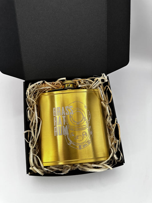 A gold brass flask engraved with 'BRASS HAT RUM' and a diver's helmet Logo, presented in a black gift box with shredded paper on a white background.
